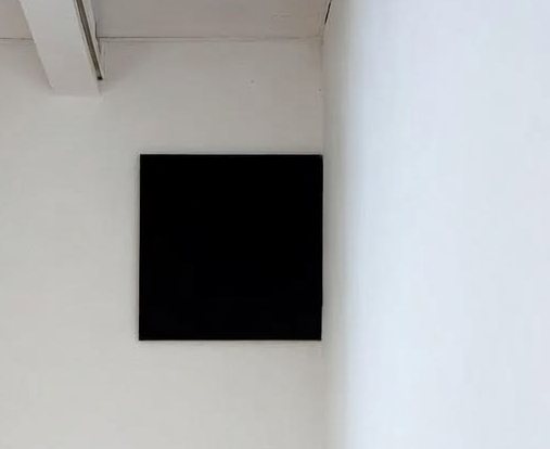 A small black square canvas hung high across the upper corner of a whitewashed room, above a bare wooden floor.