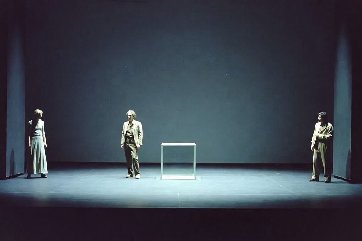 Three actors in pale suits standing far apart on a bare dark stage around a single lit glass box.