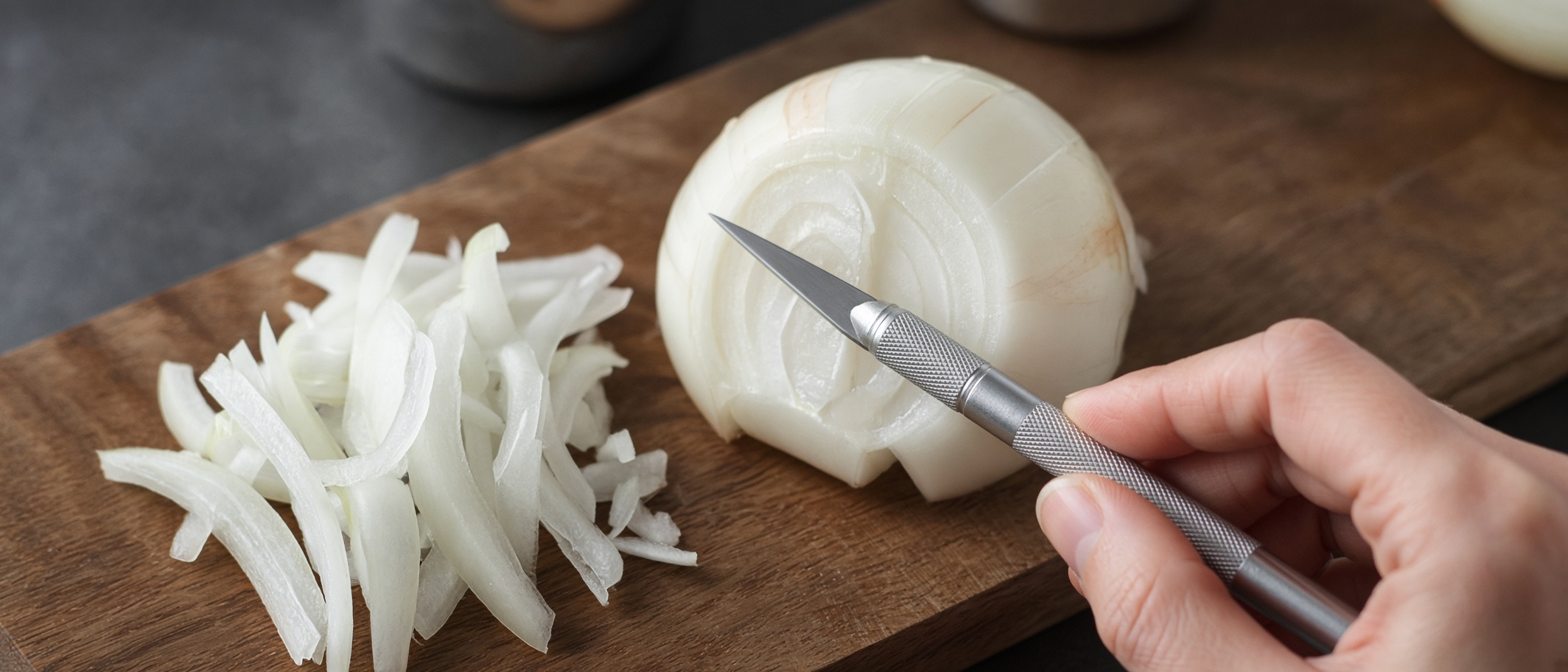 A scalpel blade drawn through a halved onion on a wooden board, the sliced pile beside it